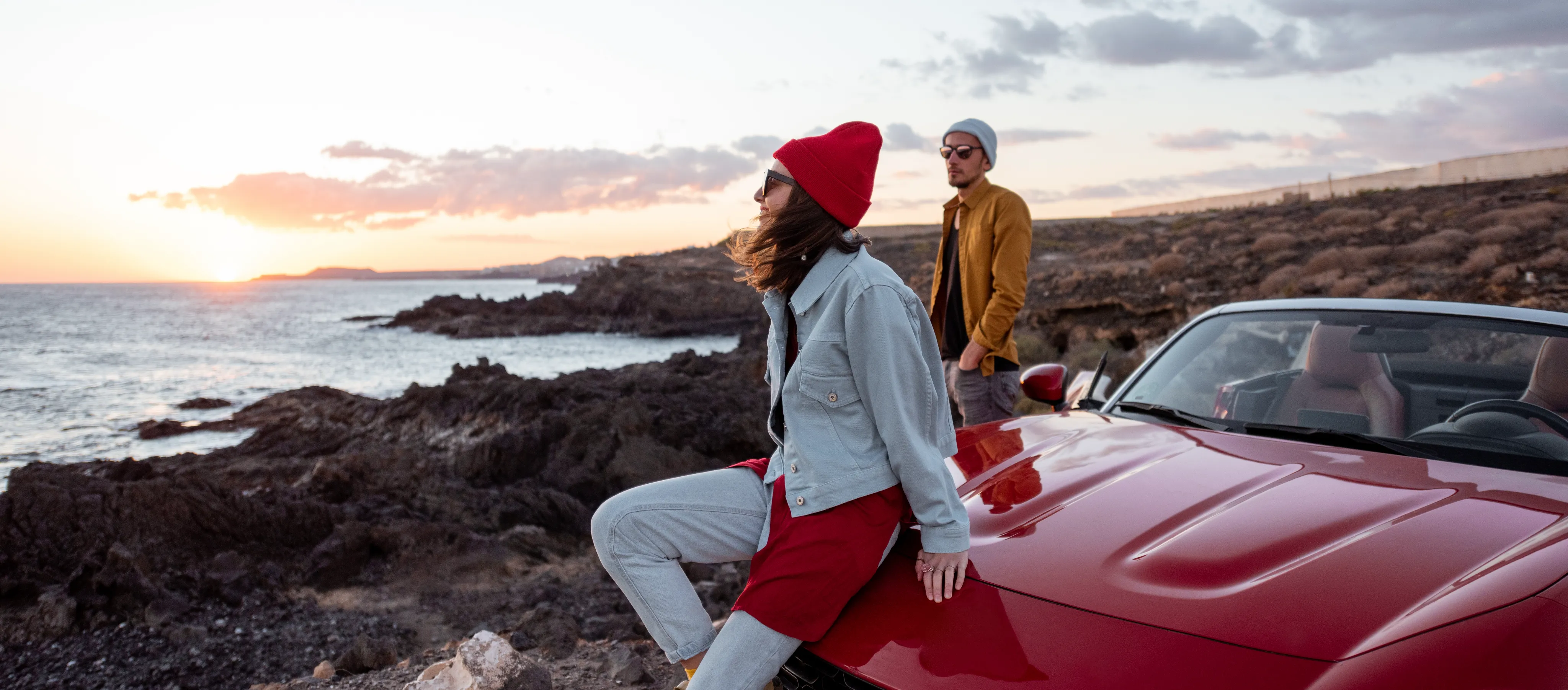 A coastal road at dusk, seen from behind two travellers looking out over the water